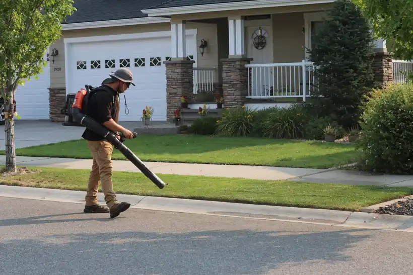A crew member blowing grass clippings off a sidewalk after a mow, with a house behind