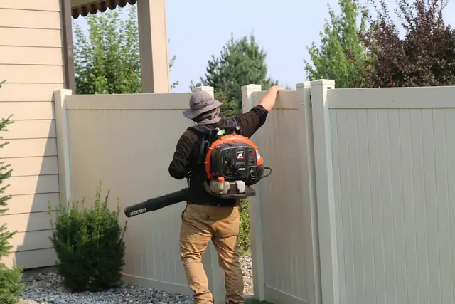 A crew member with a backpack blower clearing debris along a tan vinyl fence