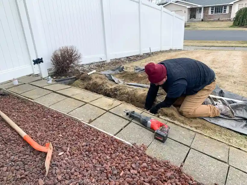 A technician kneeling at a cut sprinkler line beside pavers, with a saw and shovel