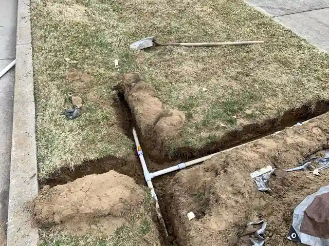 An open trench in a lawn with a new white PVC tee fitted into a sprinkler line